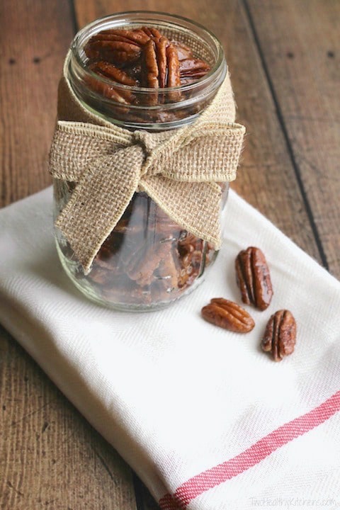 Candied pecans in a mason jar with a burlap bow. there are 3 pecans outside the jar on a napkin.